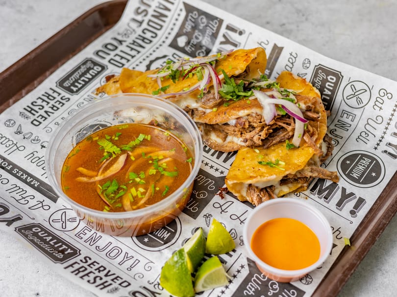 Three birria tacos with melted cheese, cilantro, and red onion, served with consommé, lime, and sauce.