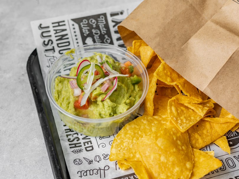 Guacamole with radish and jalapeño slices, alongside tortilla chips in a brown paper bag.