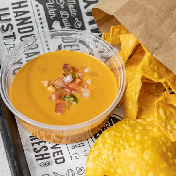 A clear plastic cup of orange queso dip with pico de gallo, next to a brown paper bag of tortilla chips.