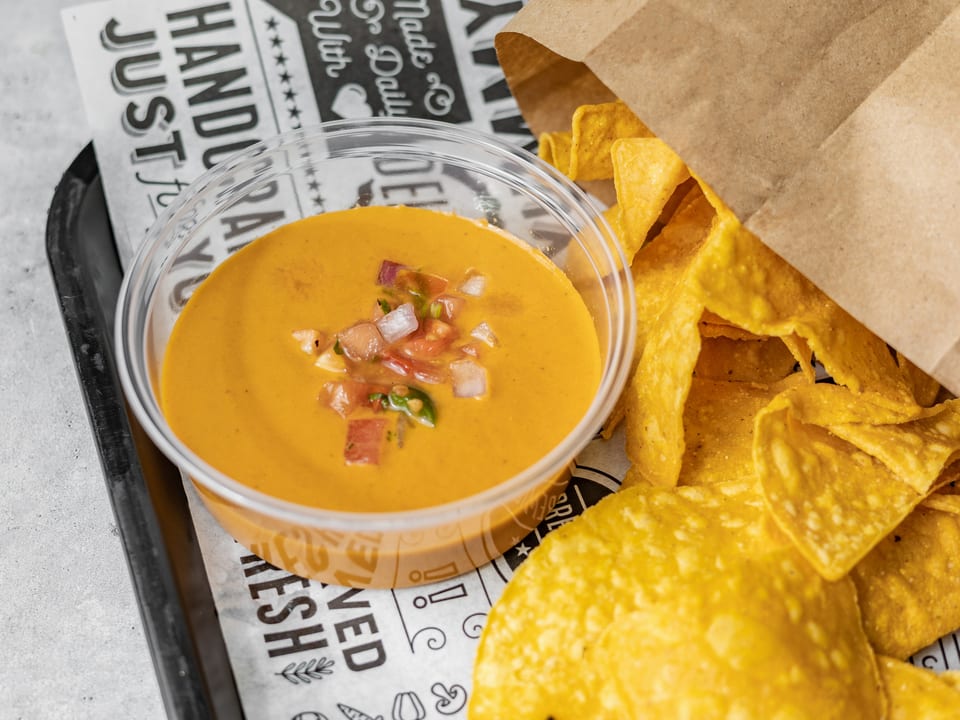 A clear plastic cup of queso dip with diced tomatoes and onions, next to a bag of tortilla chips.