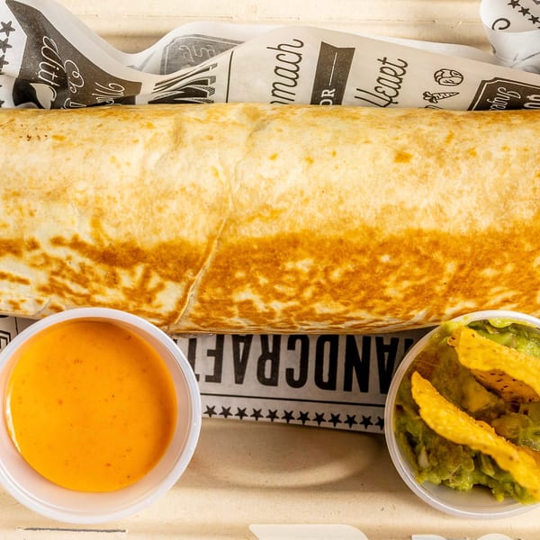 A large burrito in a paper-lined tray with sides of orange sauce and guacamole with tortilla chips.