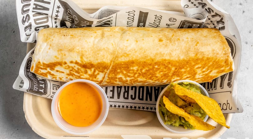 Large burrito in a paper-lined tray with sides of orange sauce and guacamole with chips.