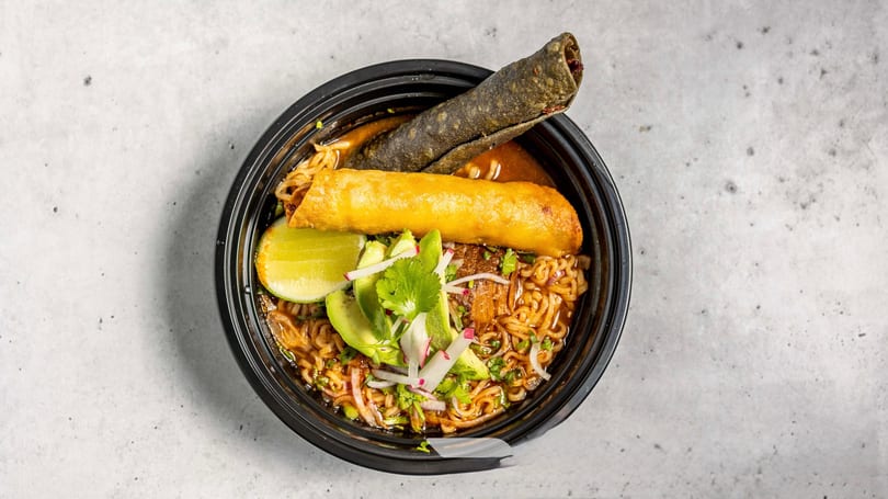 Ramen bowl with avocado, lime, cilantro, radishes, and two taquitos in a black container.